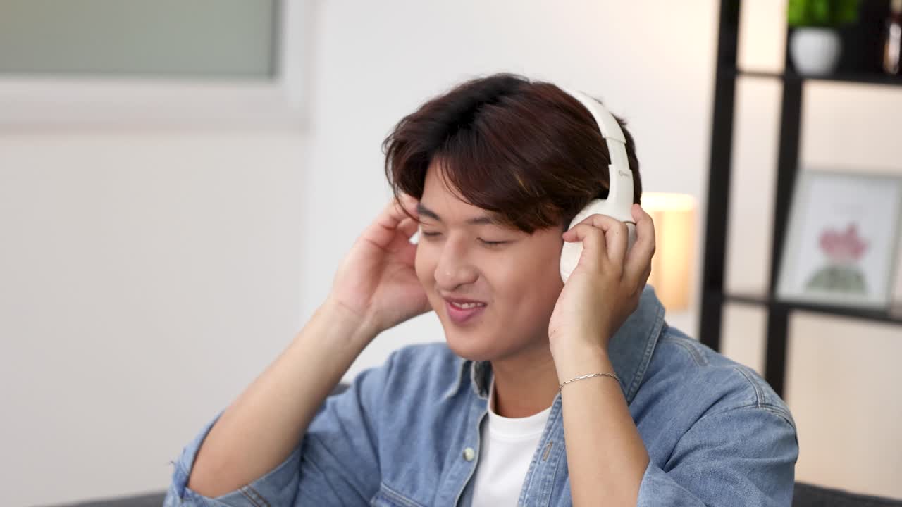 A young Asian man sits on a sofa, enjoying music through white wireless headphones indoors