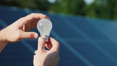 las manos del hombre sostienen una bombilla contra el fondo de los paneles solares
