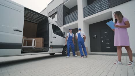 two young workers of removal company unload boxes and furniture from minibus into customer's home