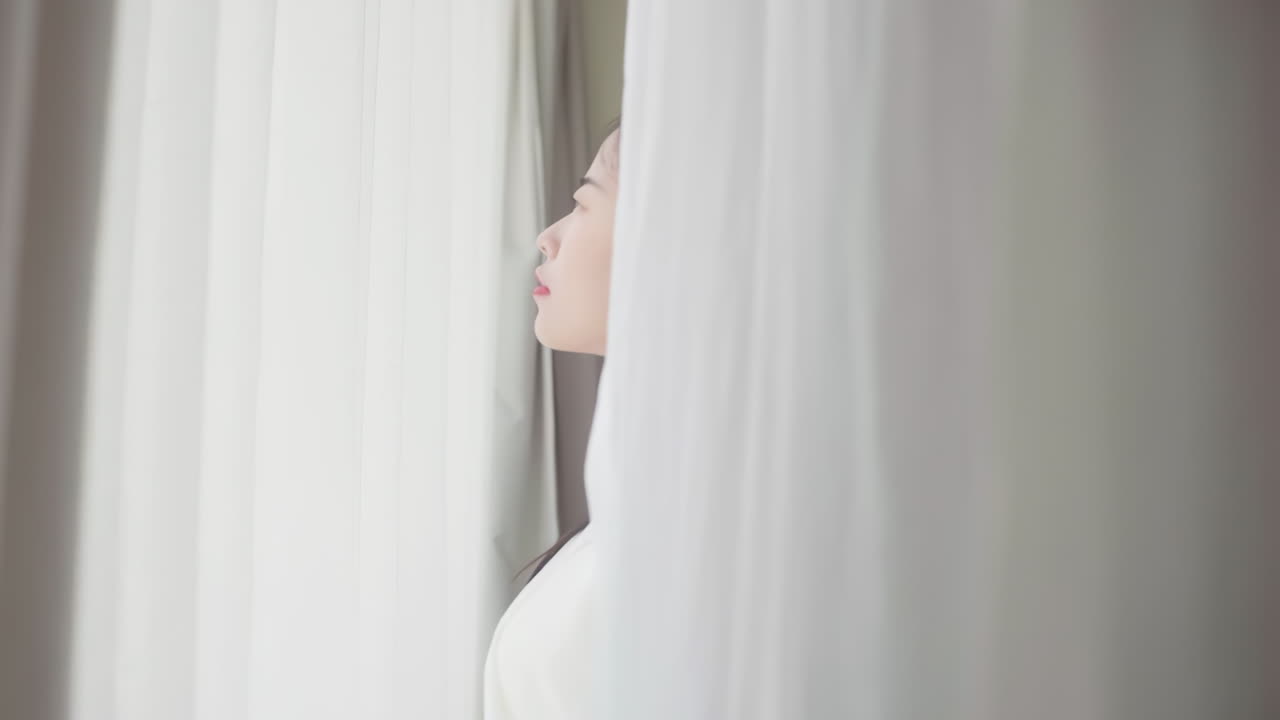 피자마를 입고 일어나는 아름다운 소녀의 실제, 큰 창문 근처에 앉아있는 침대에서 깨어나는, 완전히 휴식을 취하고, 아침에 신선한 공기와 빛을 얻기 위해 커튼을 펴는.