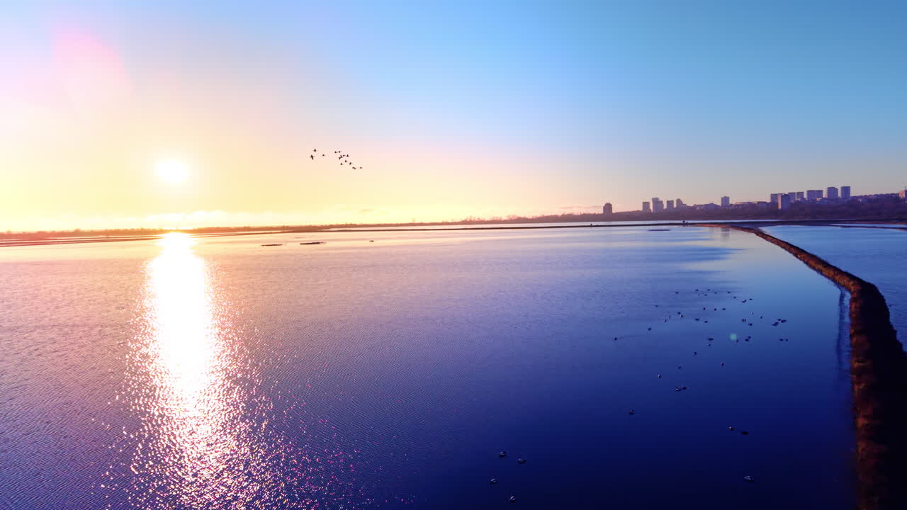 밝은 햇빛 아래 멀리 도시를 향해 뻗어 있는 염전과 제방의 기하학적 라인이에요. 새들이 있는 밝은 파란색 호수, 그림 같은 석양, 항공 뷰입니다.