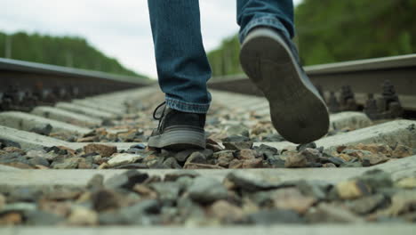 nahaufnahme der beine eines mannes, in jeans und leinwandschuhen, der auf unregelmäßiger weise auf felsigen eisenbahnschienen geht, mit einem verschwommenen blick auf grüne bäume und elektrische pfähle