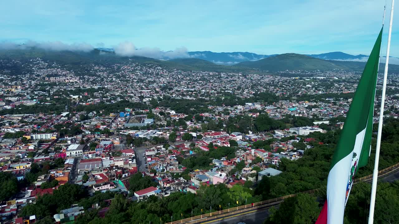 Drone aerial landscape of flag on pole waving and monument state with mountain city Oaxaca and cars traffic on highway Guelaguetza Mexico Central America travel tourism