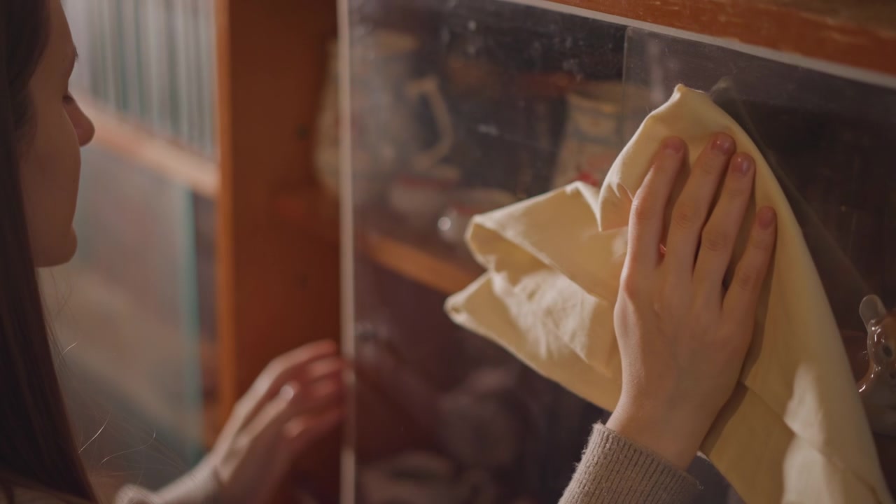 Caucasian woman wiping glass cabinet. Slow domestic cleaning scene, delicate motion removing dust from display shelves, visible reflections of collectibles, soft sweater sleeve, intimate preserved