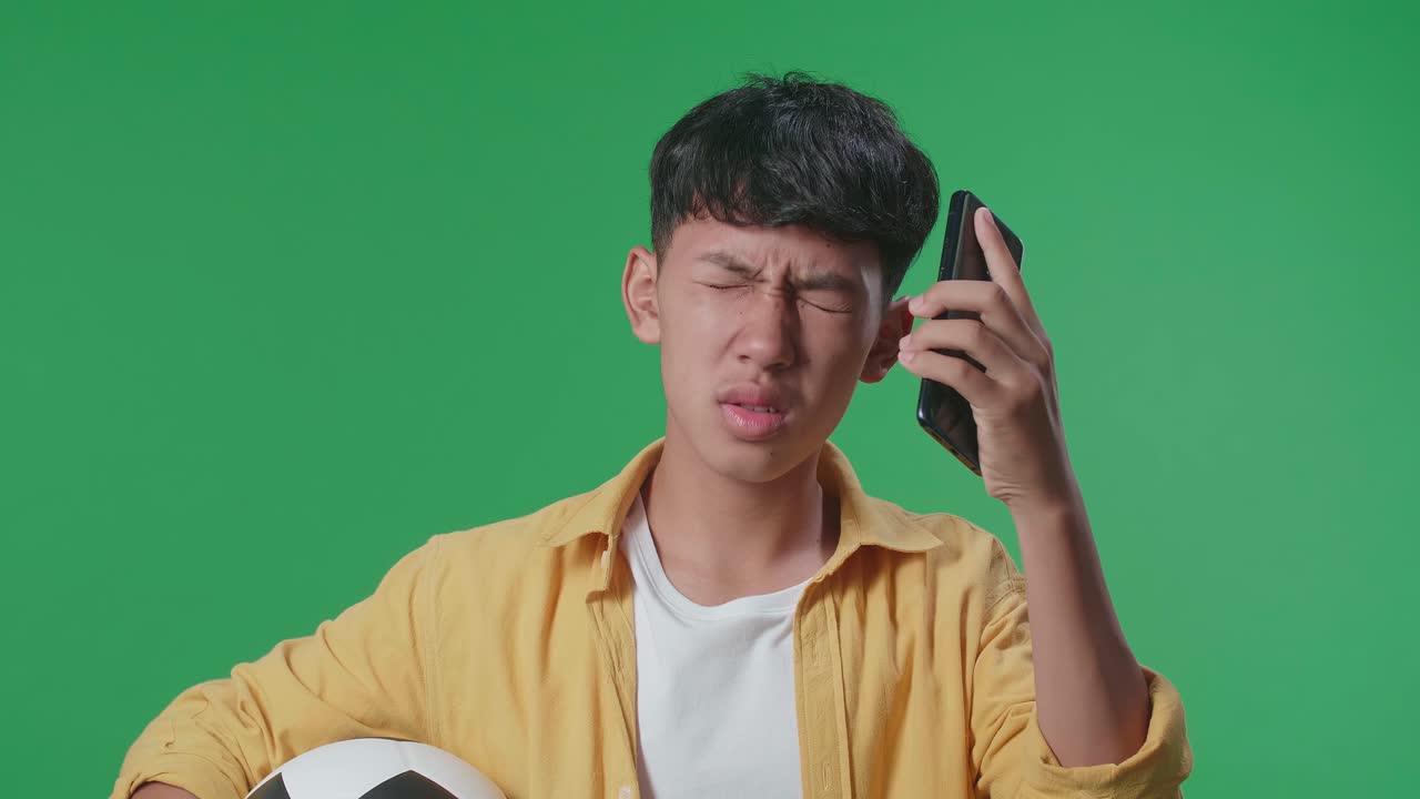 공을 들고 있는 아시아 소년은 초록색 스크린 스튜디오에서 스마트폰으로 축구를 응원하는 것을 후회하고 있습니다.