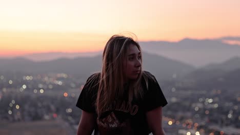 Wunderschönes-Junges-Latina-Mädchen,-Das-Vor-Einem-Bergsonnenuntergang-Am-Mount-Rubidoux-Im-Flussufer-Von-Kalifornien-Sitzt,-Mit-Rosa--Und-Orangetönen-Und-Verschwommenen-Stadtlichtern-Im-Hintergrund