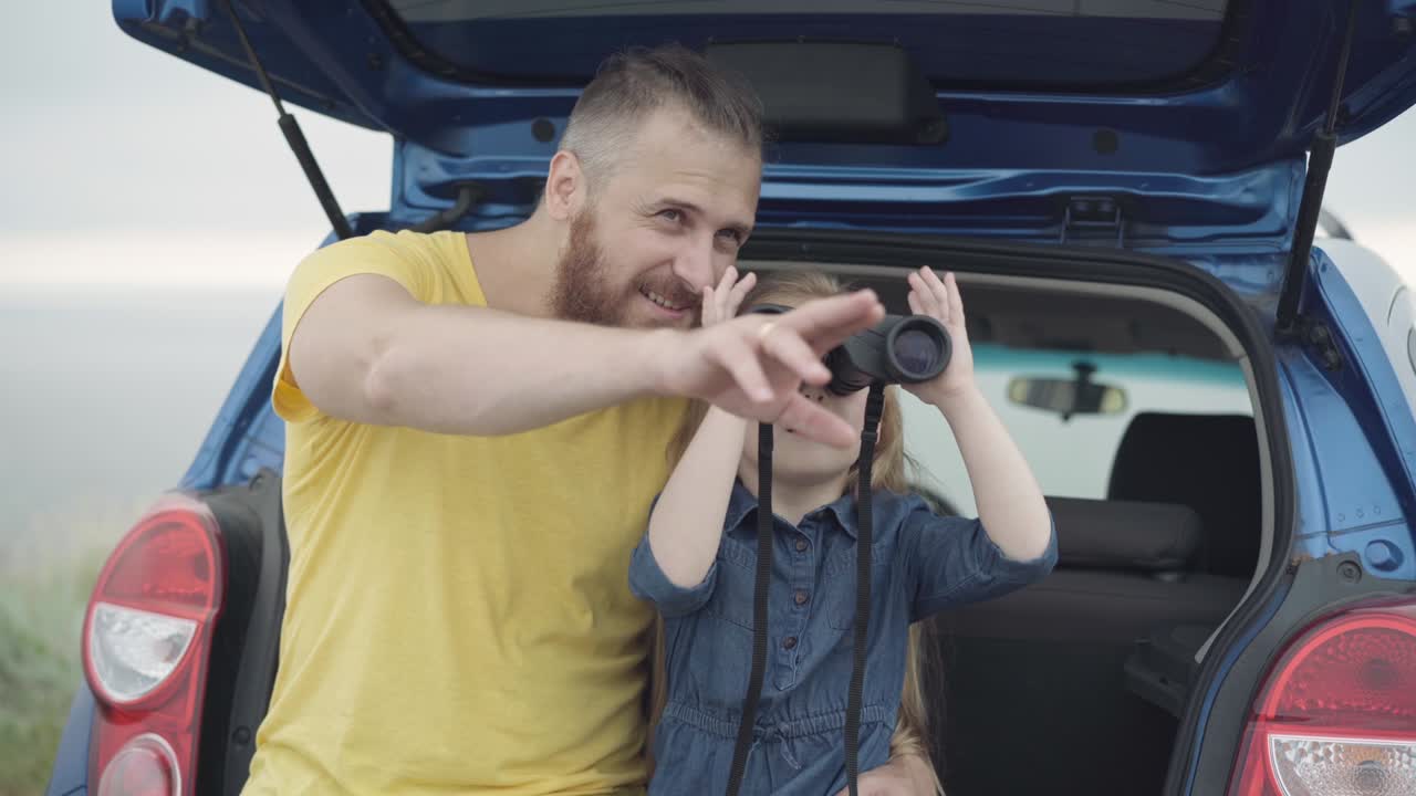 retrato de la emocionada hija caucásica usando binoculares sentada en el maletero del coche con el padre hablando. niña disfrutando del fin de semana con los padres admirando la naturaleza al aire libre. ocio familiar y unidad.