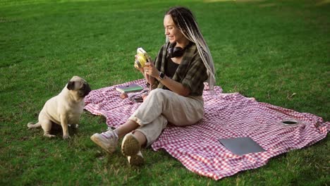 Smiling-girl-taking-photo-with-cute-pug-puppy-in-green-city-park-holding-camera,-sitting-on-plaid
