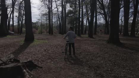 Man-walking-in-a-park-with-his-bike-stops-to-look-at-something