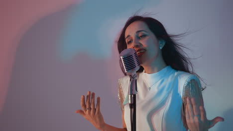 una cantante con un vestido blanco canta apasionadamente, con las manos en movimiento, frente a un micrófono vintage contra un fondo blanco. su cara expresiva y el cabello fluido añaden a la emoción