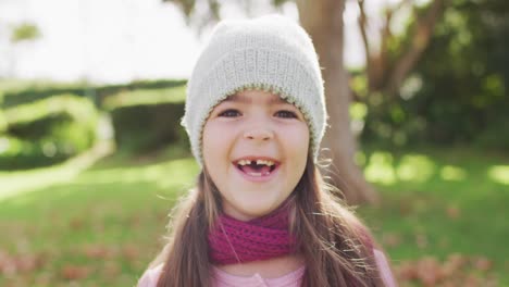 video portrait of happy caucasian girl with missing tooth wearing woolly hat and scarf in garden