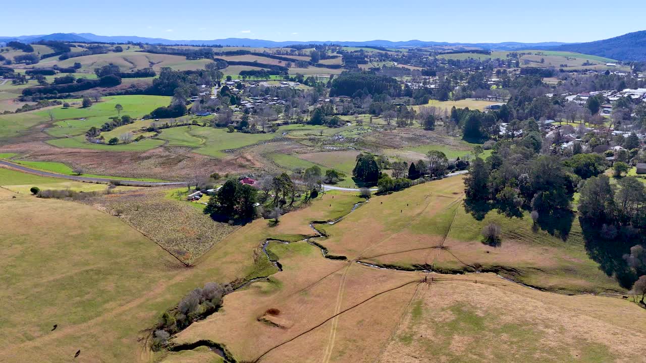Drone footage glides above sunlit rural hills, patchwork fields, and scattered trees near Dorrigo, Australia, revealing expansive countryside under clear blue skies