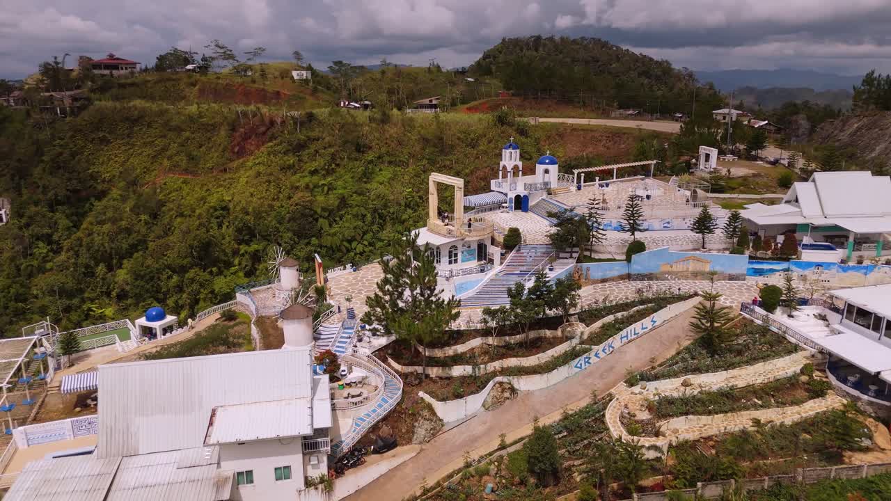 aerial shot of a hillside resort or village designed with iconic Greek Cycladic architecture—white buildings, blue domes, and pathways. Surrounded by lush, rugged mountains under a moody, cloudy sky