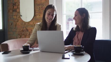 dos mujeres de negocios caucásicas sonrientes sentadas en una mesa con café, usando una computadora portátil y hablando,