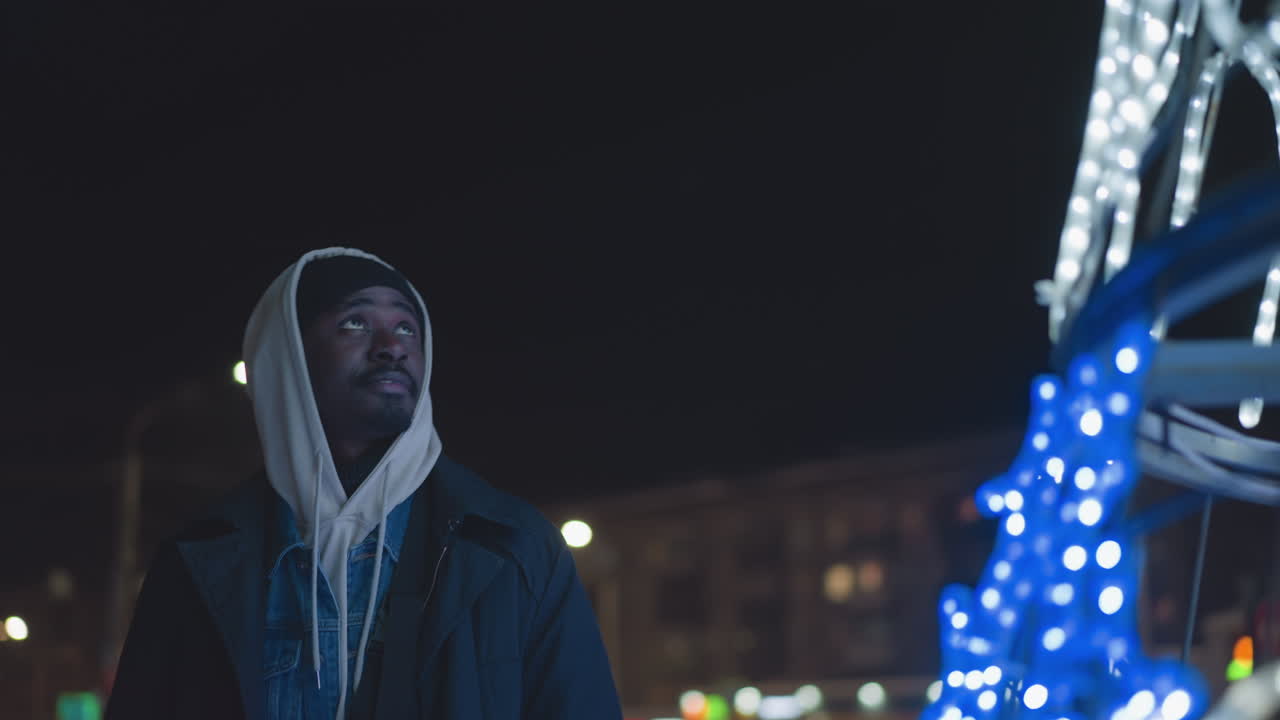 Man looking upwards at night, wearing hoodie and jacket, illuminated by street light in city setting. Modern urban backdrop with distant glowing lights, capturing calm evening vibe