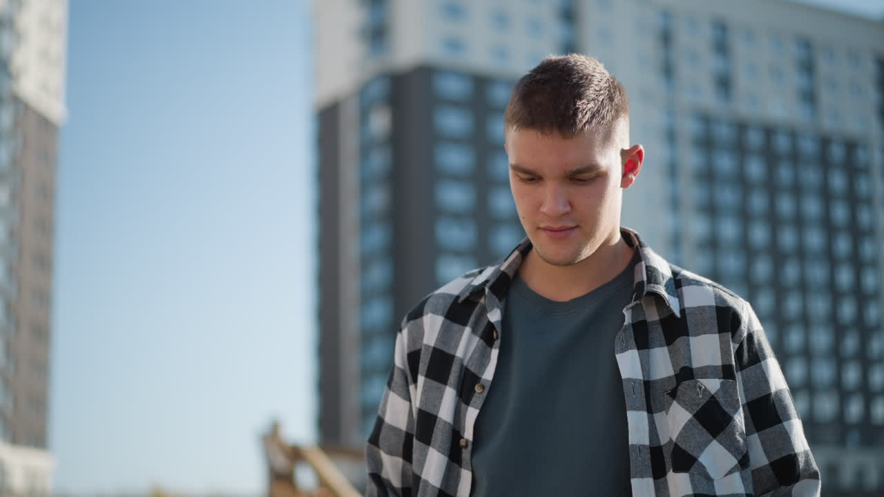 Young student in checkered shirt stands outdoors with eyes lowered in calm meditative posture as sunlight reflects off face with modern tall buildings and clear sky in background
