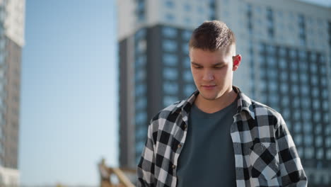 young student in checkered shirt stands outdoors with eyes lowered in calm meditative posture as sunlight reflects off face with modern tall buildings and clear sky in background