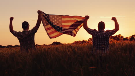 Zwei-Männer-Hissen-Glücklich-Die-Amerikanische-Flagge-über-Einem-Weizenfeld-Bei-Sonnenuntergang-4.-Juli-Konzept