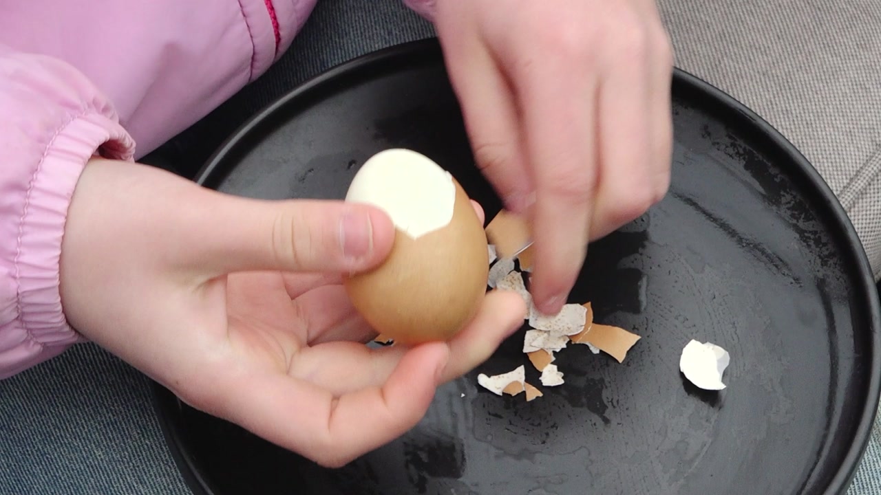 Close-up of a child’s hands peeling a boiled egg over a plate with scattered shells, with steam rising and a casual outdoor setting in the background.