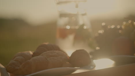 close-up of picnic setup on wooden table with fresh croissants, green grapes, orange slices, and peach under golden sunlight, refreshing water pitcher in background, with blur background