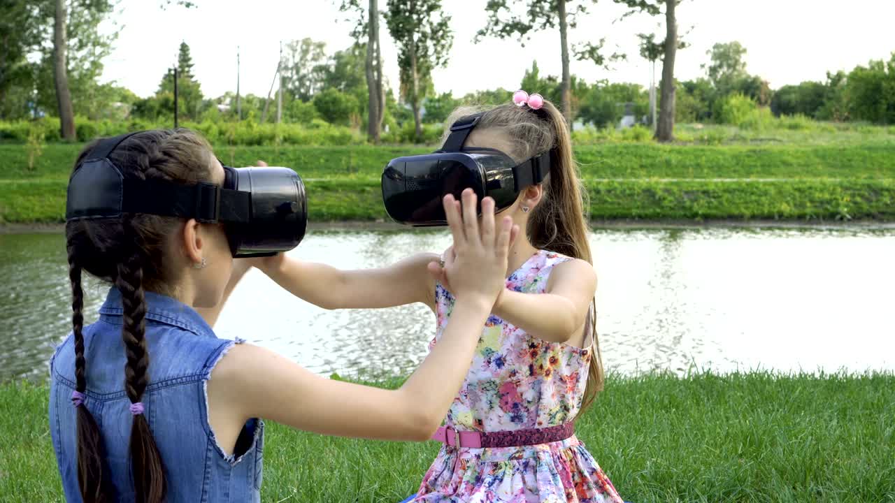 dos niñas pequeñas juegan juegos virtuales. se sientan en gafas de realidad virtual en el césped de la orilla del río en un día soleado y se toman de la mano.