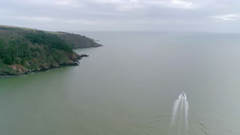 Toma-Aérea-Del-Paisaje-De-Los-Acantilados-Del-Sur-De-Devon-Con-Una-Amplia-Extensión-De-Océano-Turbio,-Y-Una-Lancha-Motora-Blanca-Que-Navega-Hacia-El-Mar-Abierto