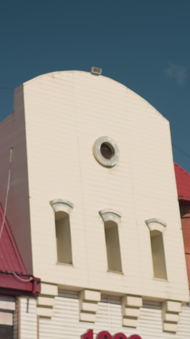 Facade of an urban building with a white central tower, circular window, and red roof under a bright blue sky, architectural details highlight modern design in a serene cityscape setting