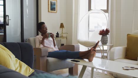 Happy-african-american-woman-sitting-on-armchair-in-living-room,-drinking-coffee