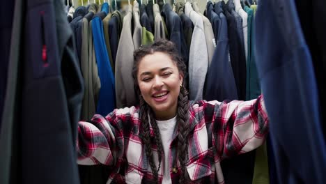 Young-black-woman-peeking-out-between-clothes-hanging-in-rail,-smiling-at-camera