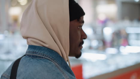 close shot of male walking in busy mall wearing beige hoodie denim jacket and beanie under hood backpack strap visible against blurred store lights and modern interior creating reflective mood
