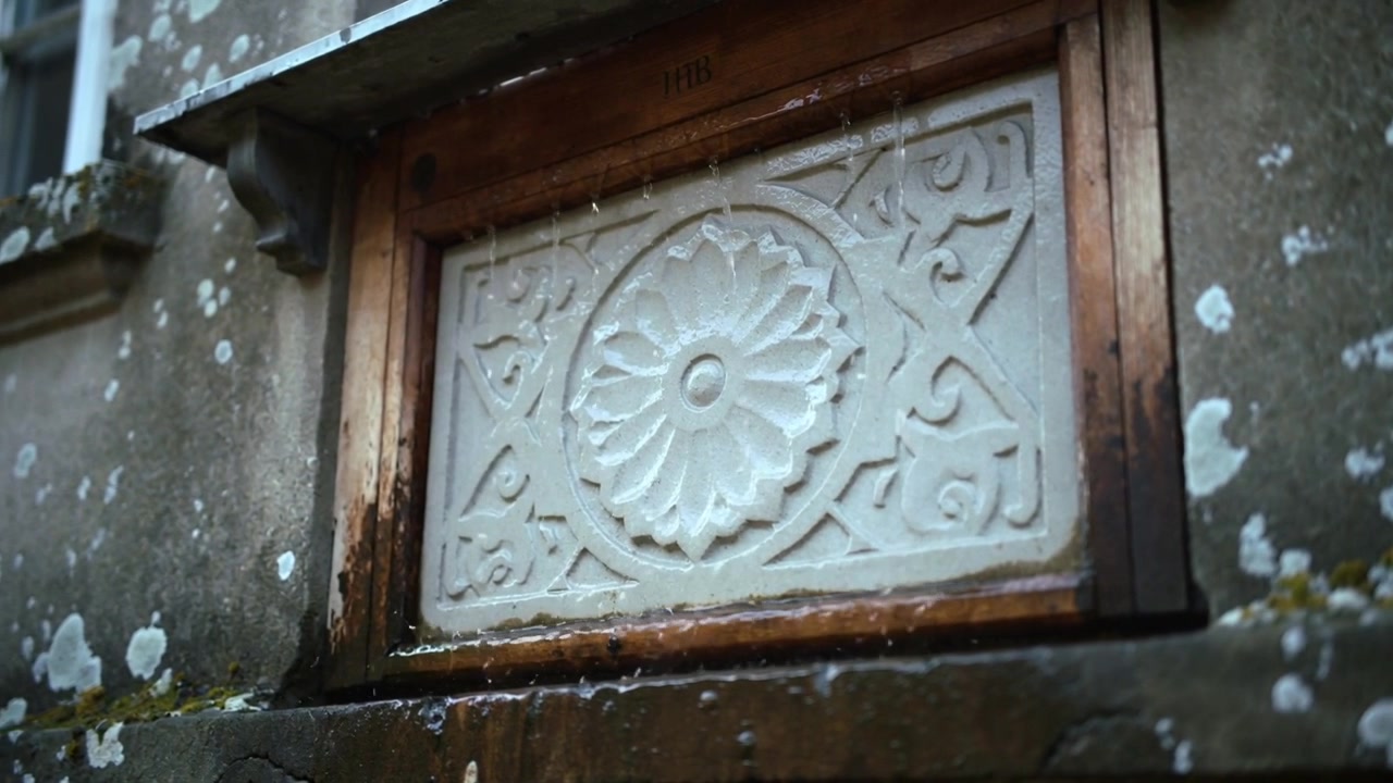 Sporgenza metallica gocciolante a causa della pioggia che manda rivoli su un pannello di pietra floreale sul muro, bagnando il muschio