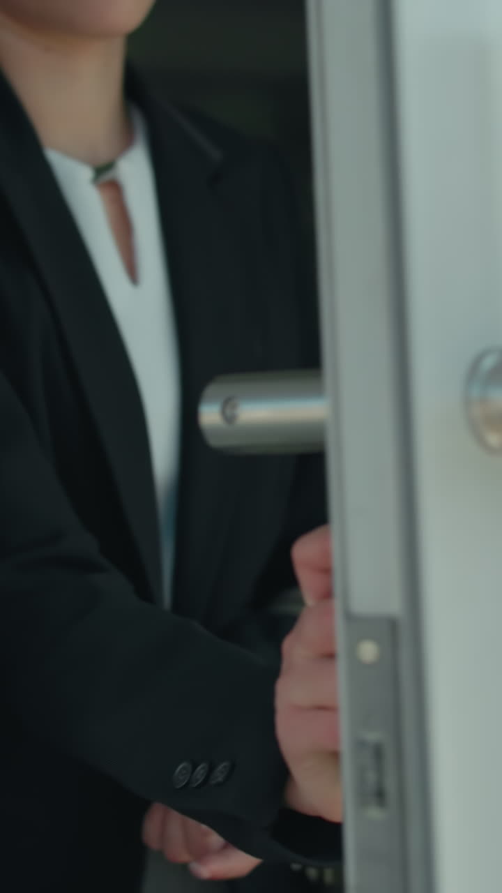 Close up of woman holding bag dressed in black suit walking out of building through glass door with urban street view in background, reflections of people and cars on glass