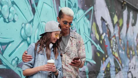 hipster couple in stylish clothes watching something on smartphone near a grafitti wall in the street