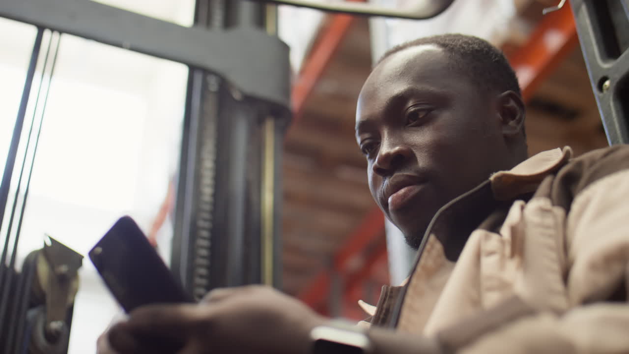 A man in a warehouse using a smartphone while operating a forklift.