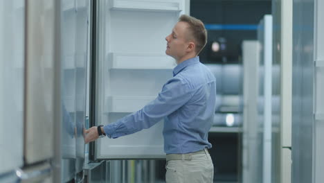 a young man in a blue shirt to open the door of the refrigerator in the appliances store and compare with other models to buy the new house