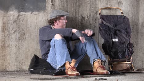 young homeless man smoking a cigarette sitting on the sidewalk under an overpass