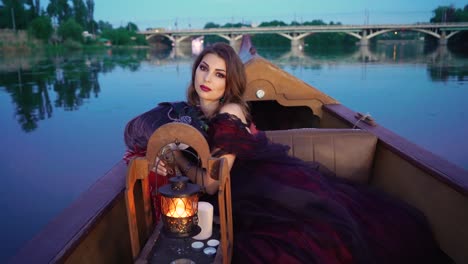 lovely sexy woman in evening dress sitting in a boat and looking at camera with passion on the river with a big bridge. pretty female floating in the evening