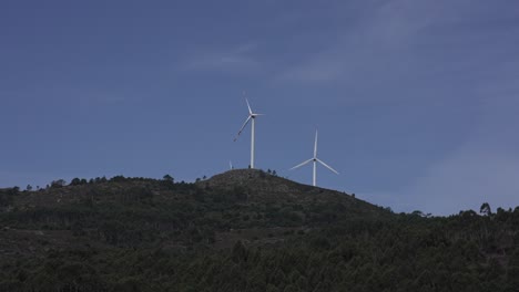 Turbinas-Eólicas-En-La-Cima-De-Una-Colina-Contra-Un-Cielo-Azul-Claro,-Rodeadas-De-Vegetación