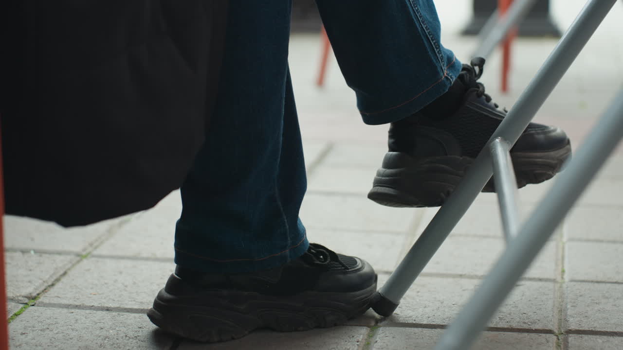 Nærbillede af sorte sko under stol, der viser denimben, der flytter fod på flisebelagt fortov ved klapbord, afslappet pendler, der venter, banker sål, ærligt bycaféøjeblik, teksturerede fliser, metalstolben