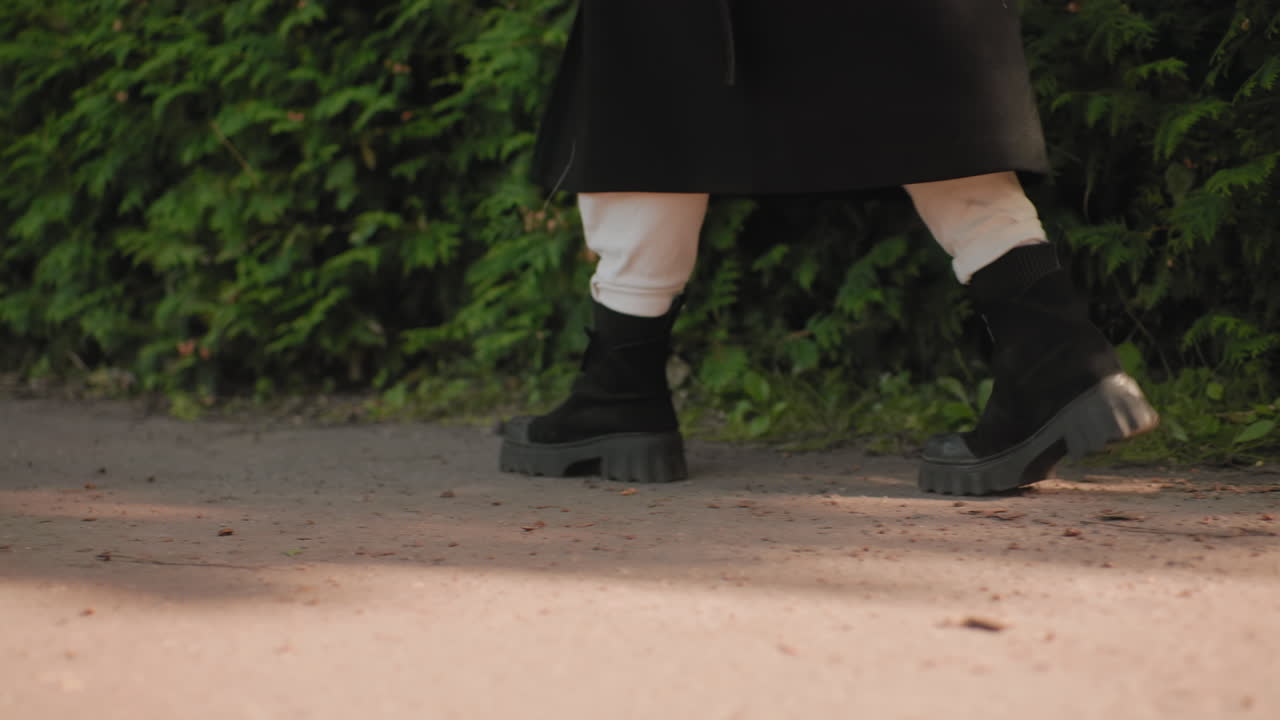 Vista de las piernas de una persona que camina en otoño con botas, pantalones blancos y un abrigo largo, paseando junto a un seto verde en un sendero iluminado por el sol, perspectiva cercana al suelo, pasos firmes, ambiente relajado, hojas crujientes bajo los pies