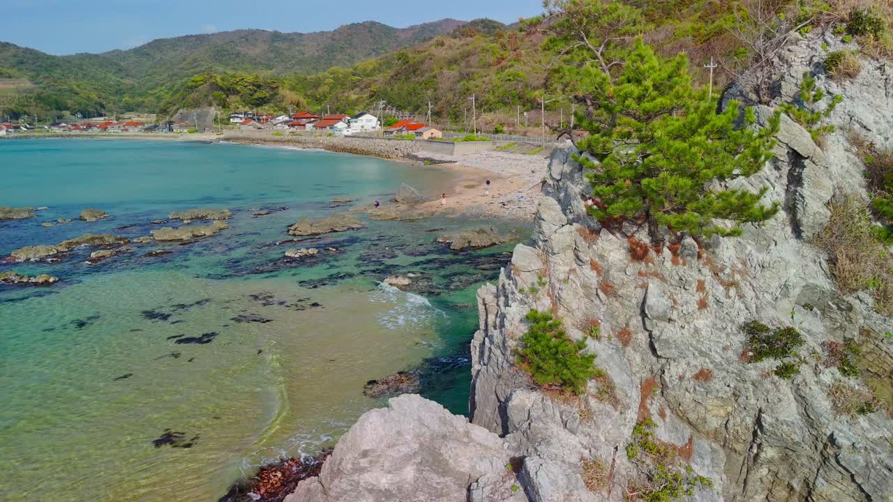 A stunning aerial shot pulls back from rugged rock formation, revealing the picturesque Uradome Coast. The clear turquoise water laps against a sandy beach where a small village is nestled at the base