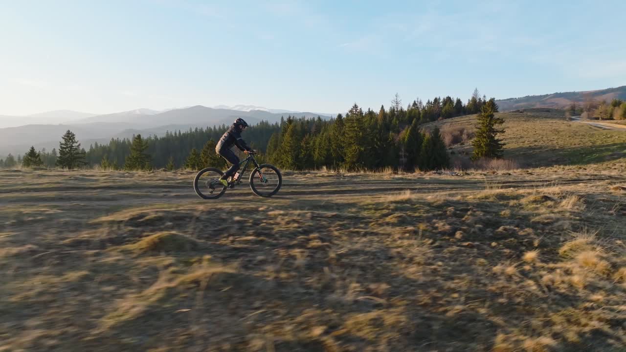 ciclista montando a lo largo de un sendero de montaña escénico al atardecer, rodeado de árboles y colinas onduladas