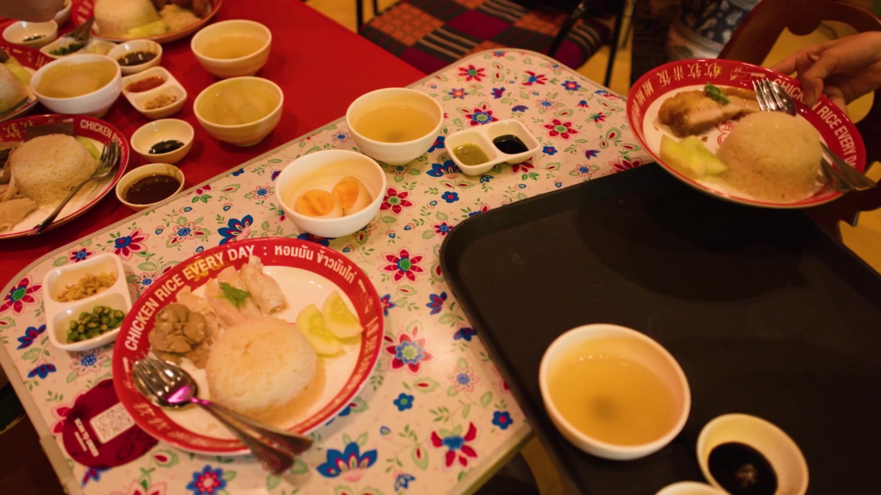 Hands arrange plates of boiled chicken, steamed rice, and soup on a floral tablecloth.