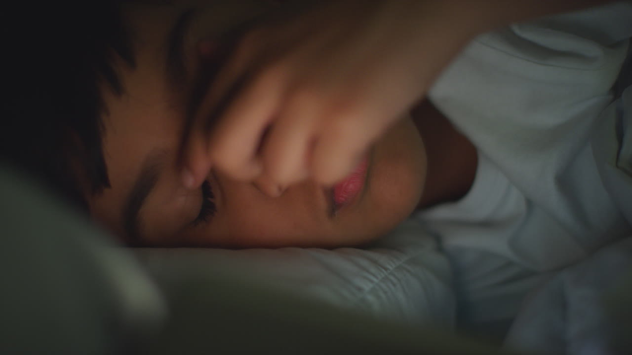 집에서 침실에서 피곤한 어린 소년이 침대에 누워서 휴대 전화를 사용하여 밤에 문자 메시지를 보내고 있습니다.