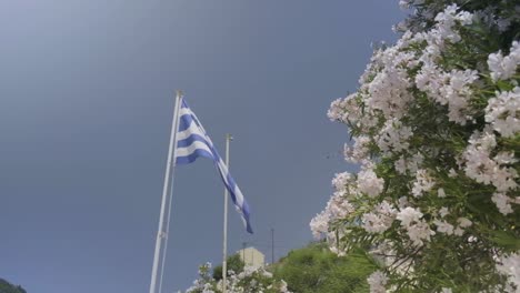 Toma-En-Cámara-Lenta-De-La-Bandera-Griega-Ondeando-En-El-Viento-Junto-A-Las-Flores-Floreciendo