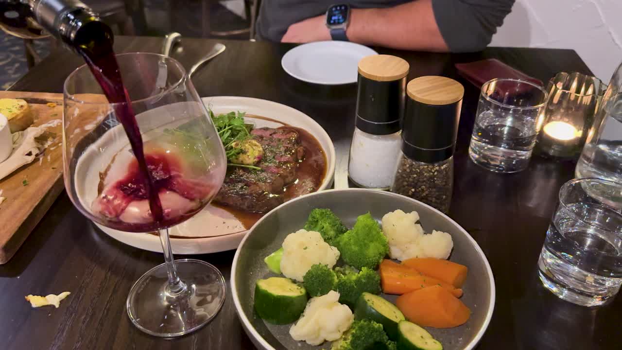 Hand pours red wine into glass beside steak, vegetables, and candlelit dinner in cozy restaurant