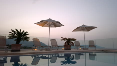 panning right shot of reflective pool at dusk with sun loungers, umbrellas - miniature palm trees with mountains in distance