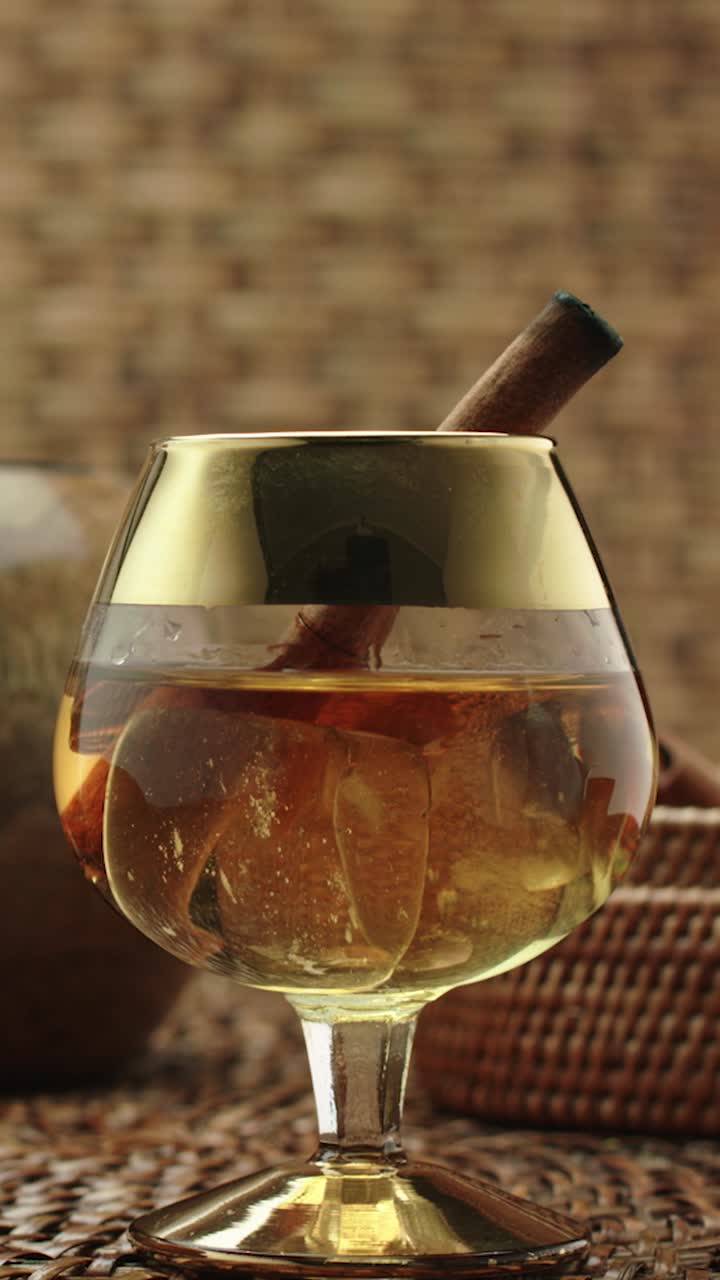 Golden spiced rum cocktail with cinnamon stick and ice cubes served in luxurious glass, panning shot