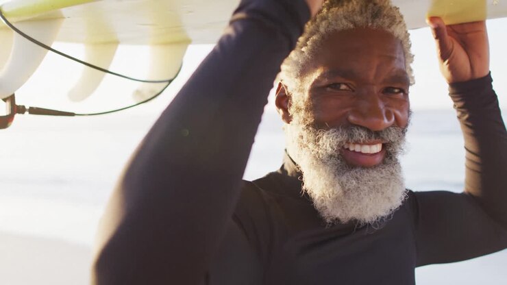 Portrait of happy senior african american man carrying surfboard on sunny beach