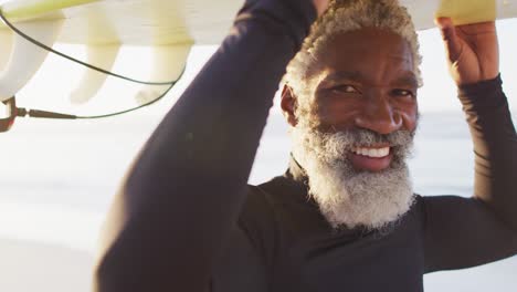 portrait of happy senior african american man carrying surfboard on sunny beach